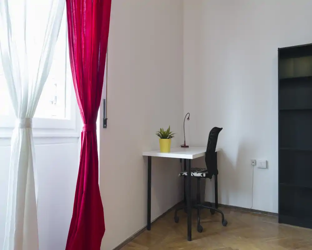 Angolo luminoso e minimale con una piccola scrivania, sedia da ufficio, mensole e tende vicino alla finestra — adatto come zona giorno o angolo studio in un appartamento in affitto.