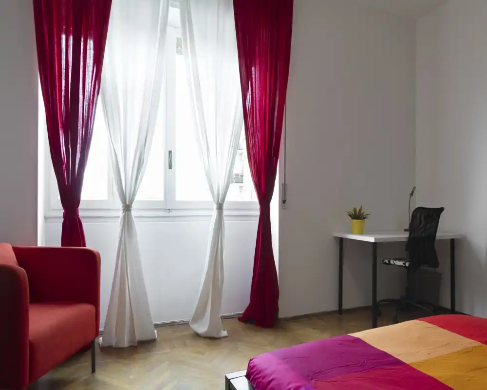 Camera da letto luminosa e colorata con letto visibile, poltrona rossa e una piccola scrivania vicino alla finestra; tende leggere filtrano la luce naturale creando un’atmosfera accogliente.