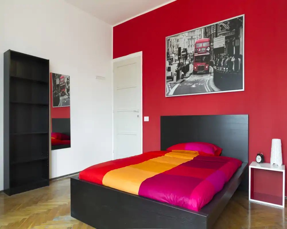 Camera da letto moderna e luminosa con struttura del letto bassa nera e copriletto colorato rosso-arancione. Parete accentuata in rosso con stampa di Londra, comodino semplice e libreria: ambiente vivace e di stile.