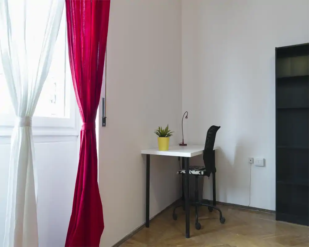 Angolo soggiorno/lavoro luminoso e minimal con scrivania bianca, sedia ergonomica, lampada e una pianta accanto a una finestra con tende rosse e bianche.