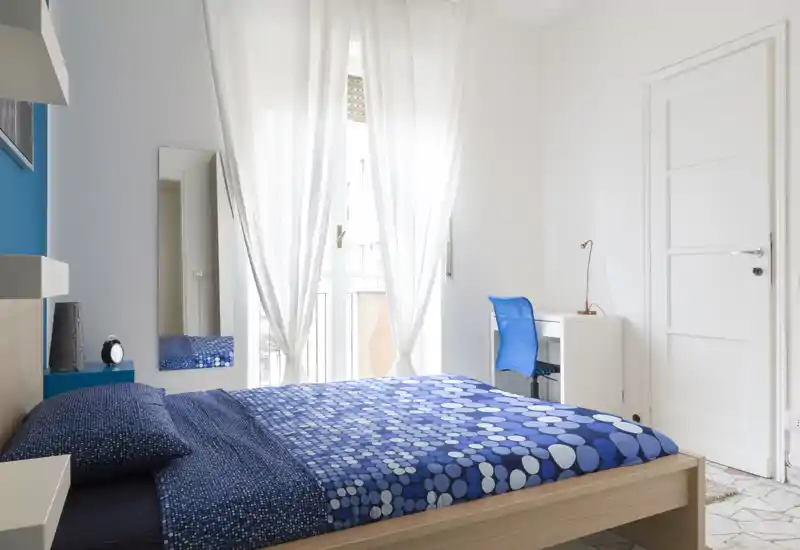 Bright, modern bedroom with a blue patterned bed, full-size wooden bed frame and a small desk by the window. Large sheer curtains let in ample natural light and the room has clean, minimal styling.