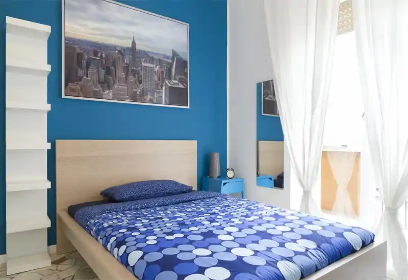 Bright, modern bedroom with a wooden bed frame, patterned blue bedding and natural light from a large window creating a fresh, welcoming atmosphere.