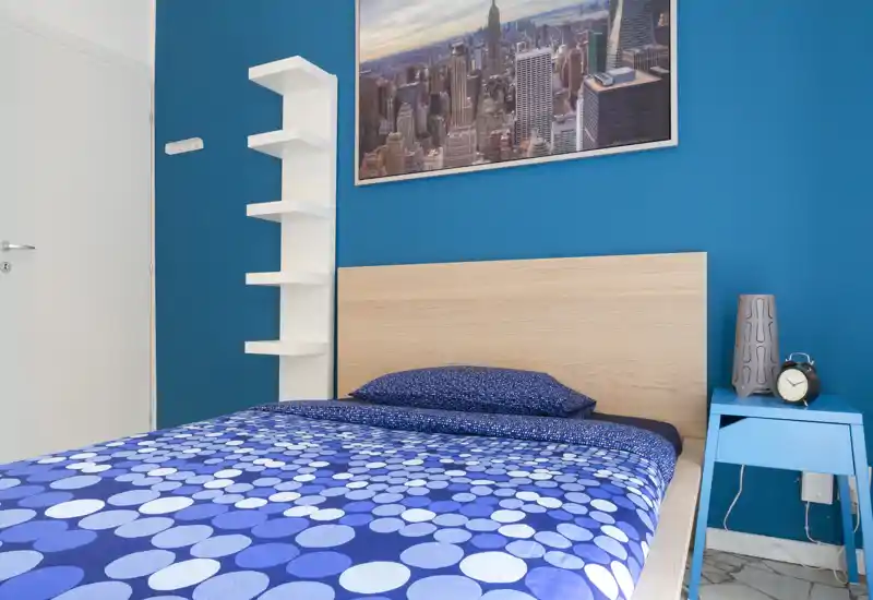 Bright, modern single bedroom with a wooden headboard, patterned blue bedding and a bedside table with an alarm clock. Clean lines, bold blue accent wall and decorative city-print artwork create a fresh, inviting space.