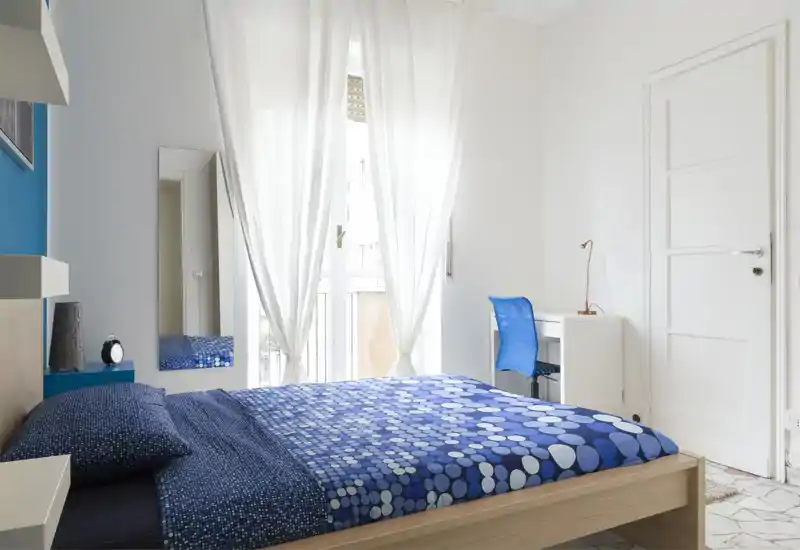 Bright, modern bedroom with a patterned blue duvet, wooden bed frame and a desk by the window. Large sheer curtains let in abundant natural light, creating a fresh and airy feel ideal for a rental listing.