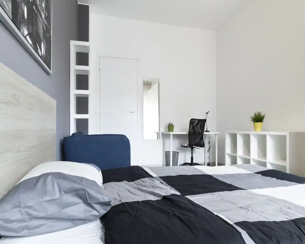 Bright, modern bedroom with a large bed in the foreground, a compact workspace and minimalist storage. Neutral tones and simple decor create a calm, welcoming atmosphere.