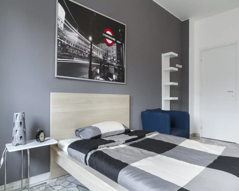 Bright, modern bedroom with a double bed dressed in monochrome bedding, a bedside table with lamp and alarm clock, a blue armchair and framed wall art above the headboard.