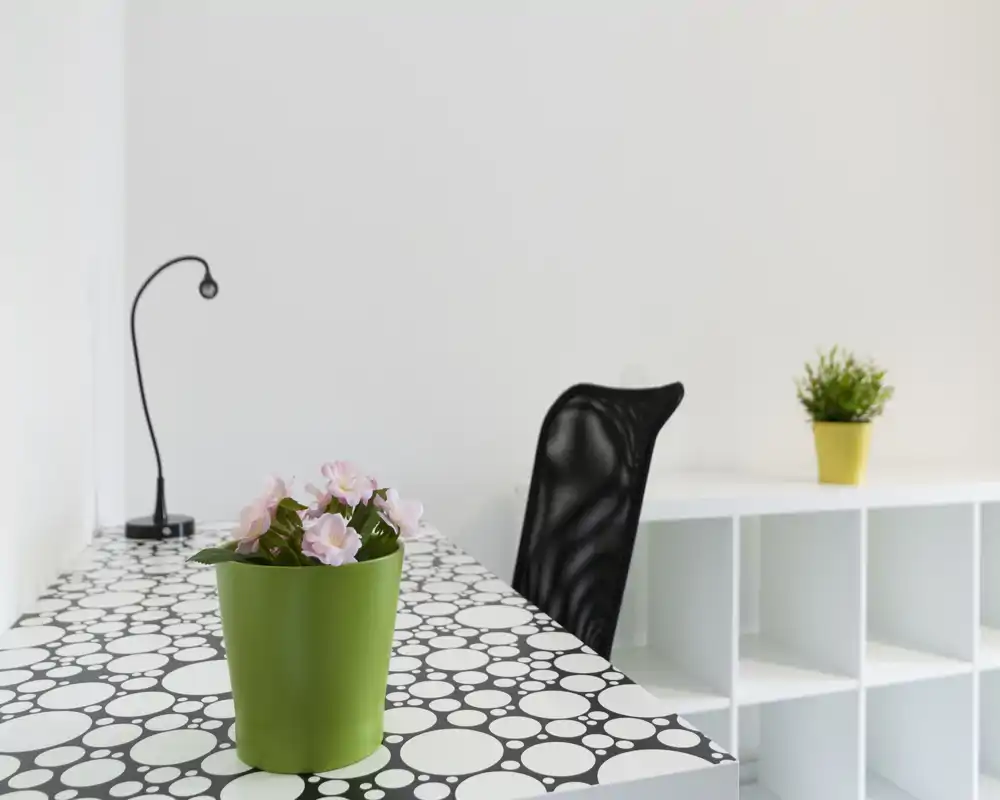 Bright, minimal workspace with a patterned desk, ergonomic black chair, small desk lamp and potted plants that add a touch of greenery.