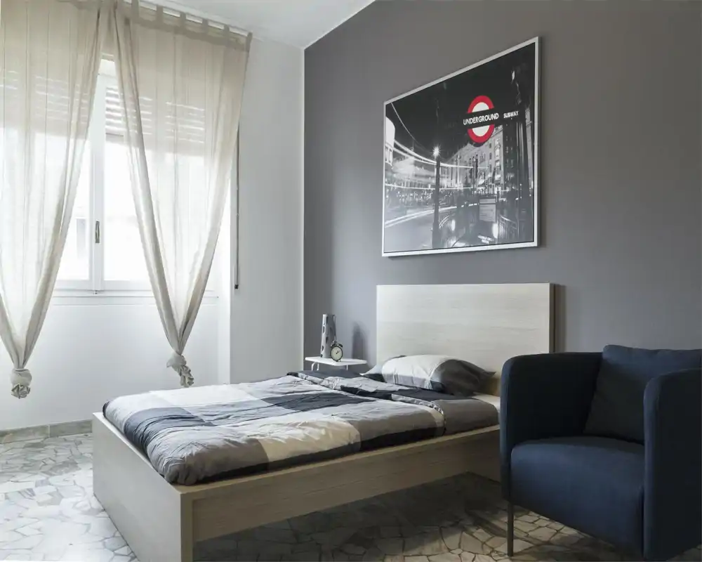 Bright, modern bedroom with a light wood bed, patterned bedding and a comfortable armchair by the window. Soft natural light, neutral tones and a framed city artwork create a calm, welcoming atmosphere ideal for listings.
