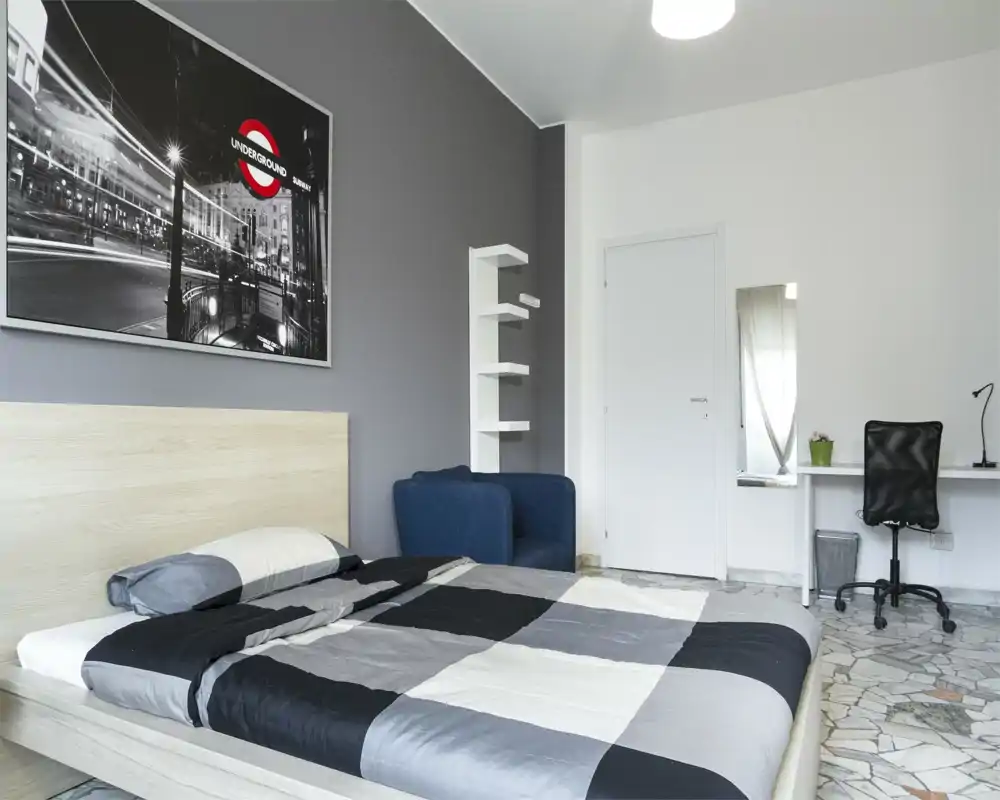 Bright, modern bedroom with a double bed dressed in gray-black bedding, a blue armchair, framed wall art and a small desk. Clean lines and neutral tones create a calm, well-styled space ideal for rental listings.