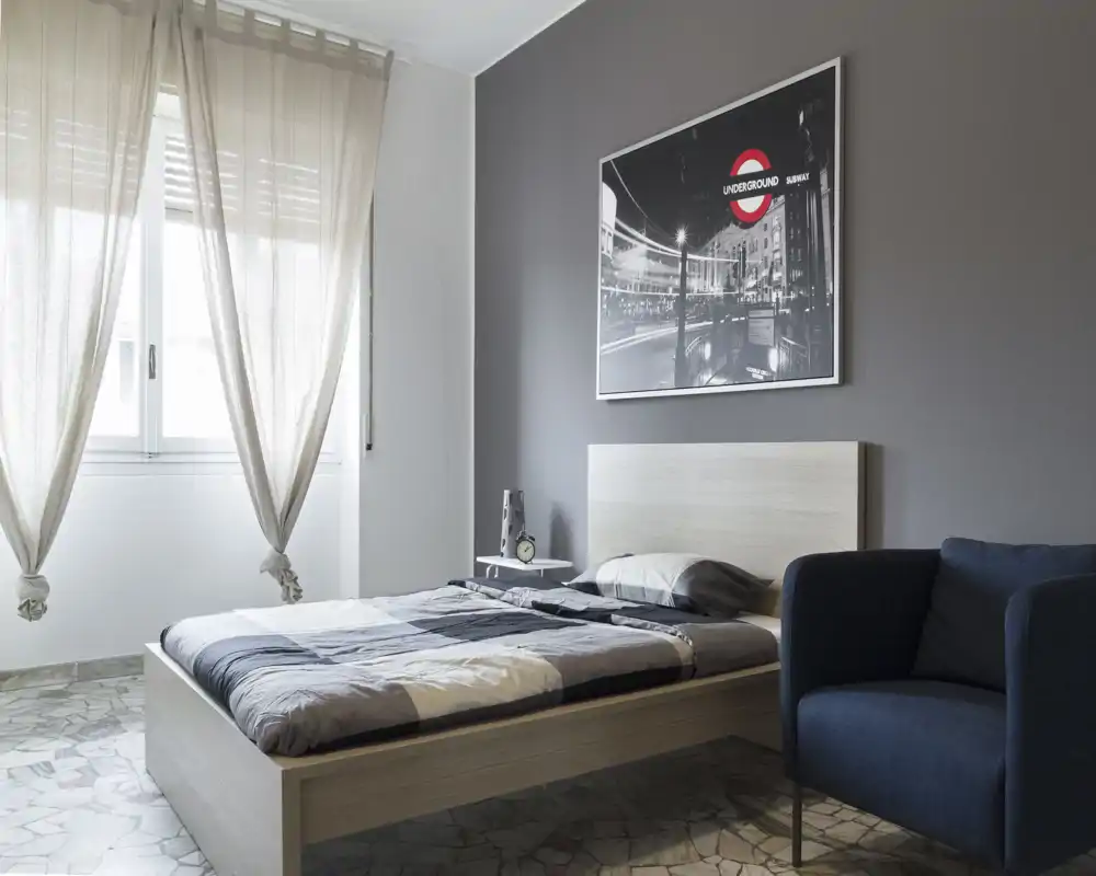 Bright, modern bedroom with a double bed, soft natural light from a large window, minimalist decor and a comfortable armchair.