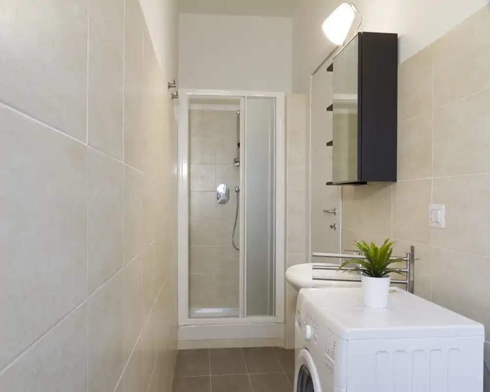 Compact modern bathroom with a walk-in shower, wall-mounted sink, mirrored cabinet and a washing machine. Neutral beige tiles and bright lighting create a clean, functional space.