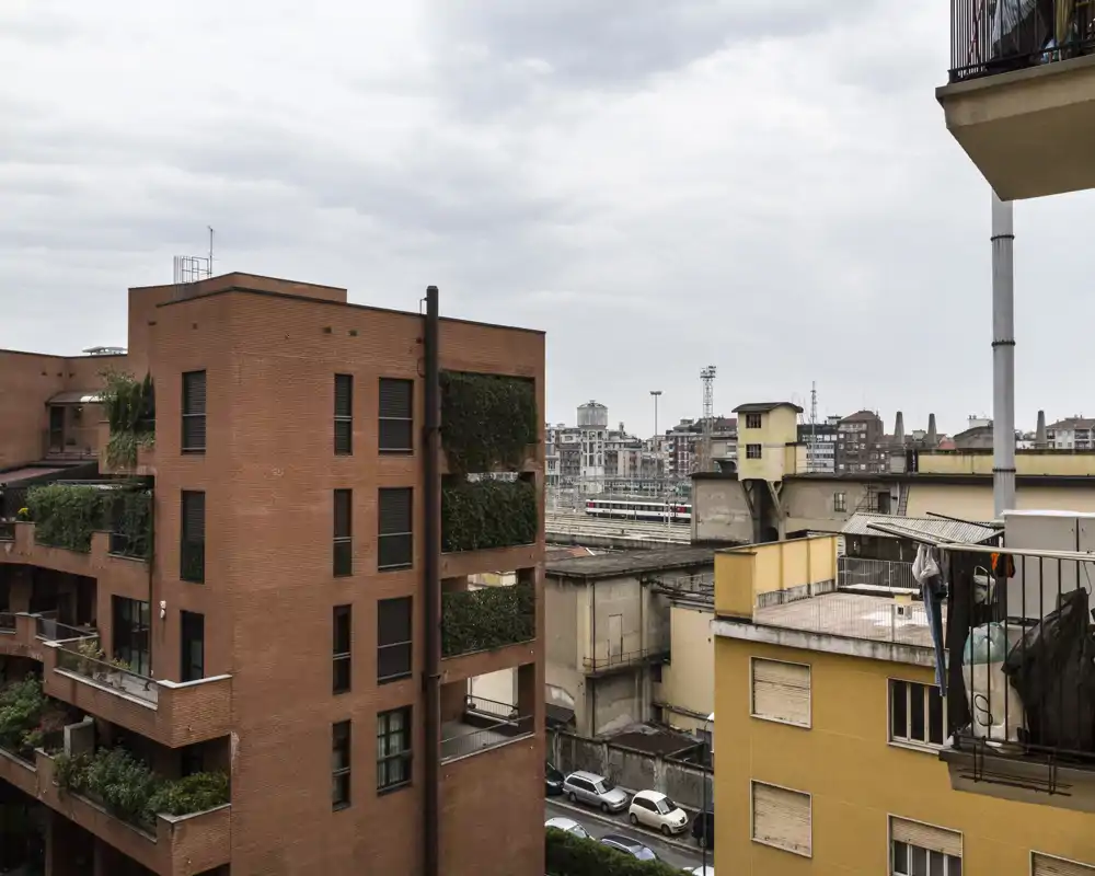 Urban outdoor view from a balcony showing neighboring brick and stucco buildings, rooftop terraces and street-level details under an overcast sky.