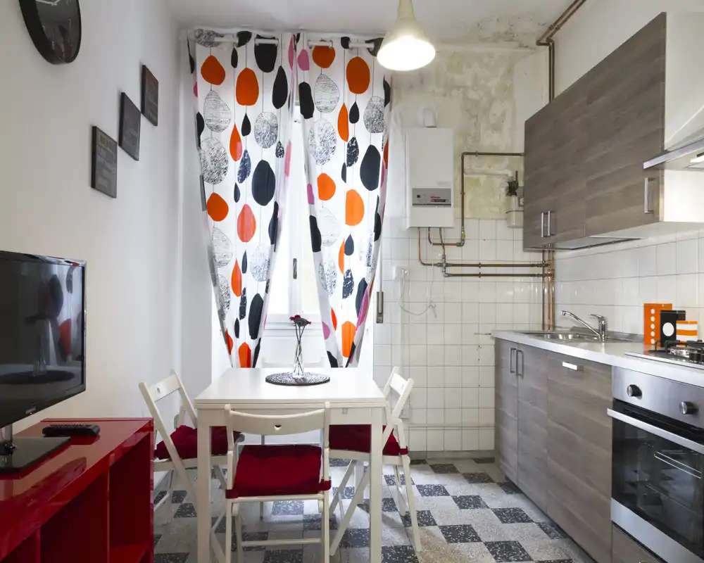 Compact kitchen with modern wood-effect cabinets, built-in oven and a small dining table by a bright window with patterned curtains — practical and inviting for short stays.