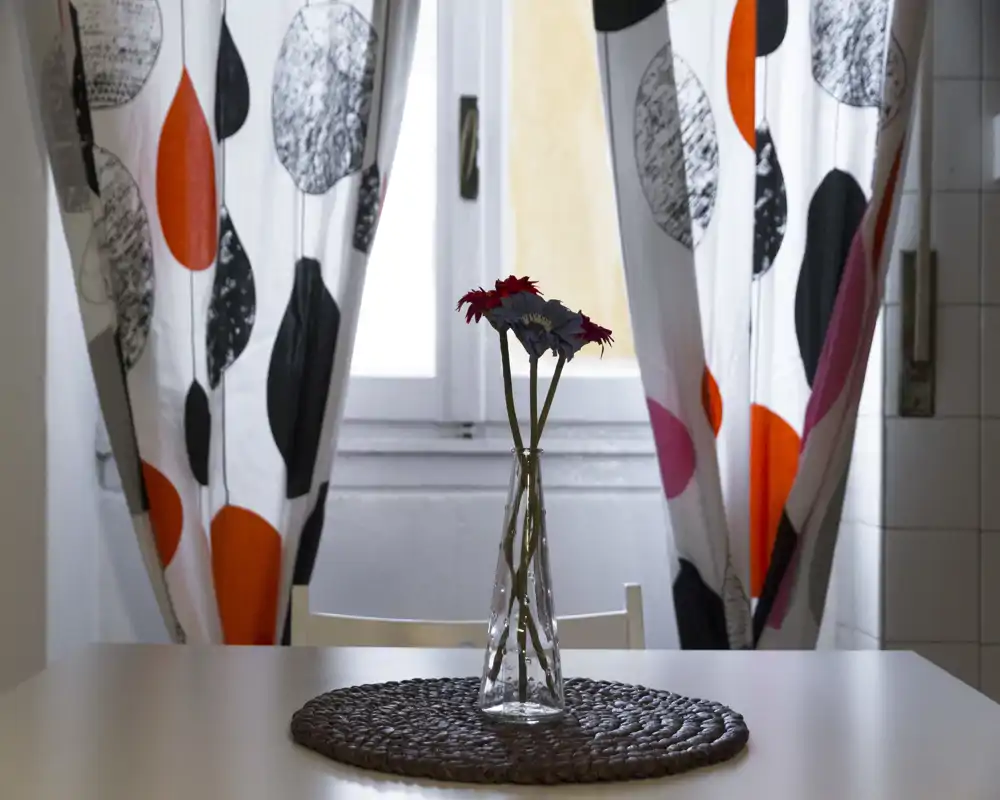 Small dining or breakfast table by a window with patterned curtains, a woven placemat and a slender glass vase holding a few decorative flowers — bright, simple and cozy for morning meals.