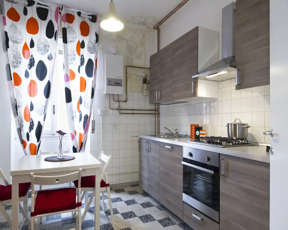Bright, compact kitchen with modern wood-effect cabinets, built-in oven and gas hob, small dining table by a window with bold patterned curtains.