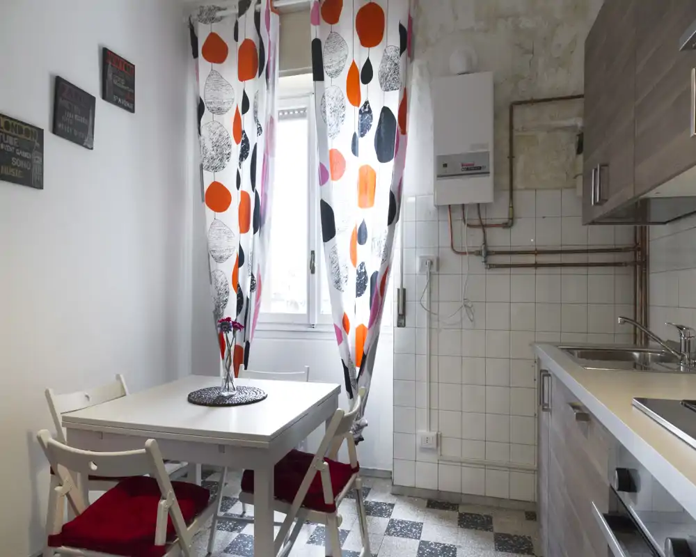 Compact, well-lit kitchen with a small dining table, four chairs with red cushions, patterned curtains and modern cabinetry — ideal for a cozy apartment.