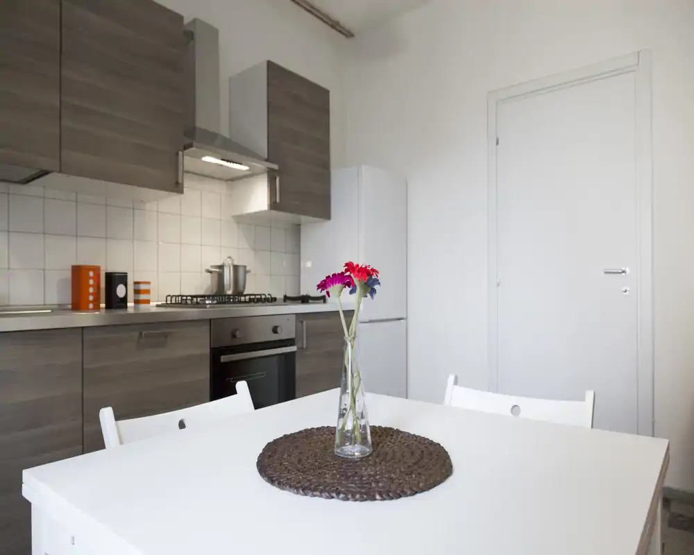 Bright modern kitchen with wooden cabinets, built-in oven and gas hob; white dining table in the foreground decorated with a vase of flowers.