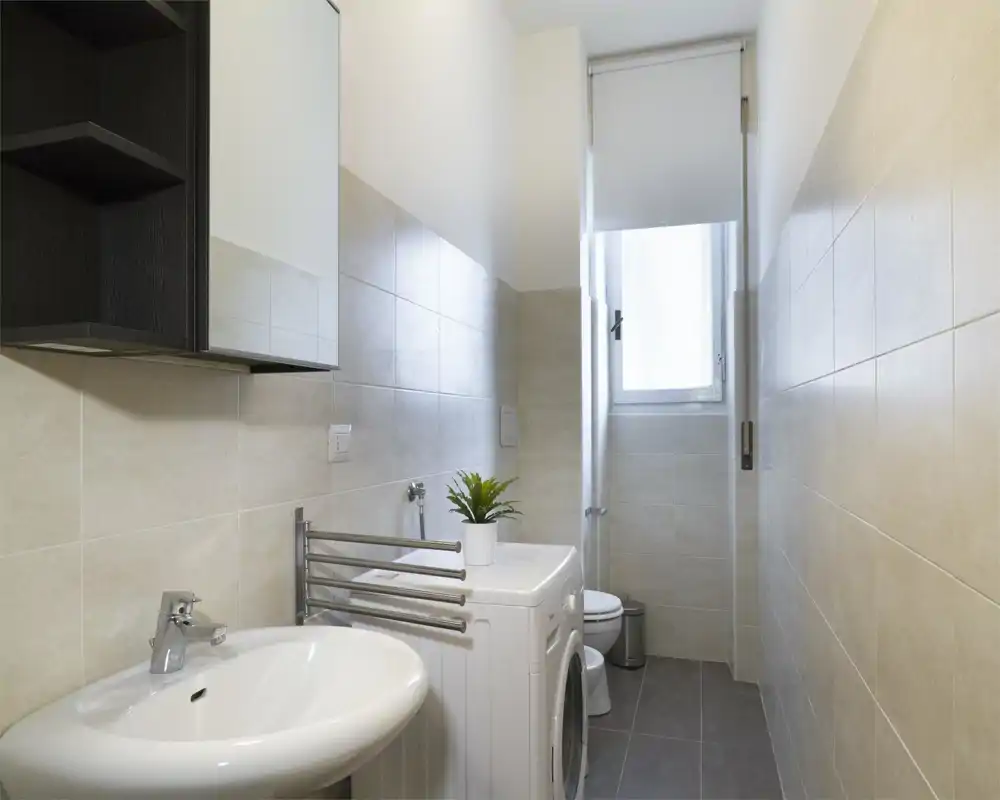 Bright, modern bathroom with wall-mounted tiles, a sink, toilet and a washing machine; neutral tones and a small potted plant add a clean, functional feel.