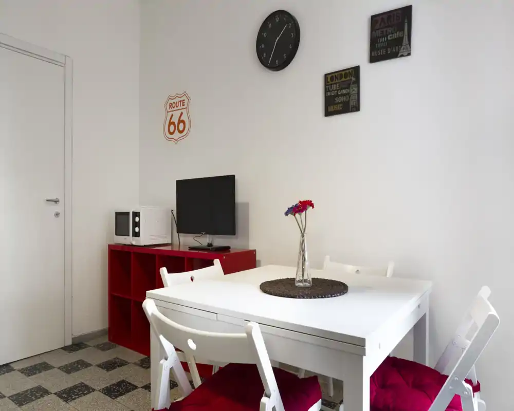 Bright and simple dining area with a white table, folding chairs with red cushions and a decorative vase. Compact layout includes a small media unit and microwave — practical for short stays.