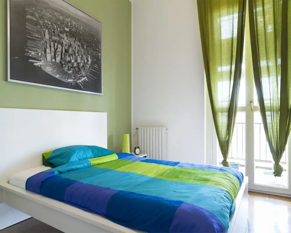 Camera da letto moderna e luminosa con letto matrimoniale colorato, testiera bianca e ampia finestra con tende verdi che lasciano entrare luce naturale.