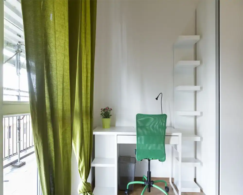 Zona giorno luminosa e minimal con una scrivania bianca contro la parete, una sedia girevole verde e una tenda verde abbinata; ideale come angolo studio compatto con luce naturale dalla finestra del balcone.