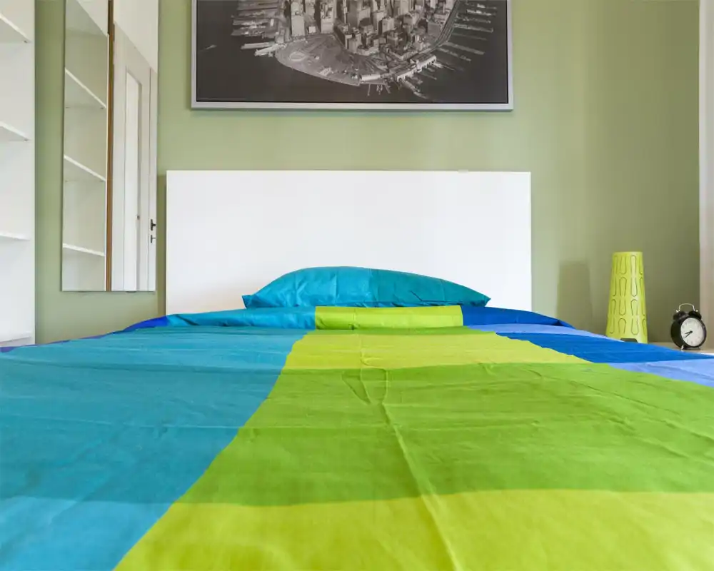 Camera da letto moderna e luminosa con testiera bianca pulita, biancheria colorata blu e verde, abat-jour e sveglia sul comodino; ben illuminata e fotografata con nitidezza.