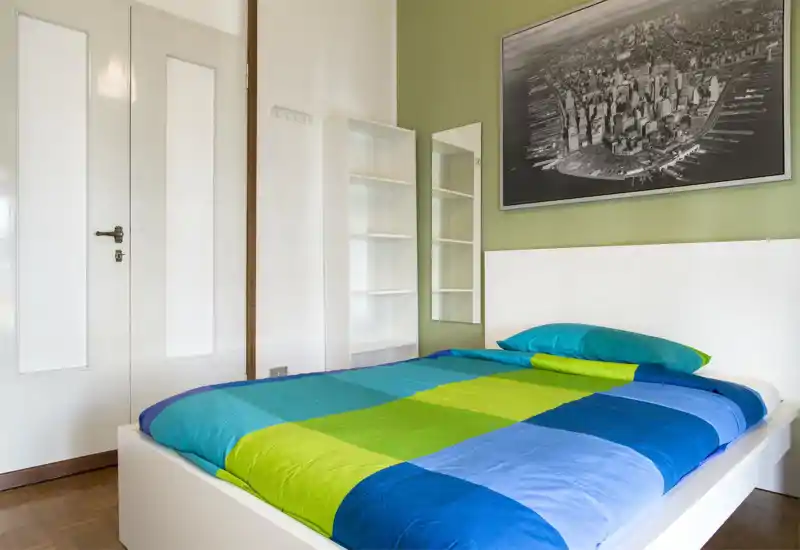 Camera da letto luminosa e moderna con un ampio letto con biancheria colorata, testata bianca, quadro e scaffali — ben illuminata e arredata in modo semplice e fresco.
