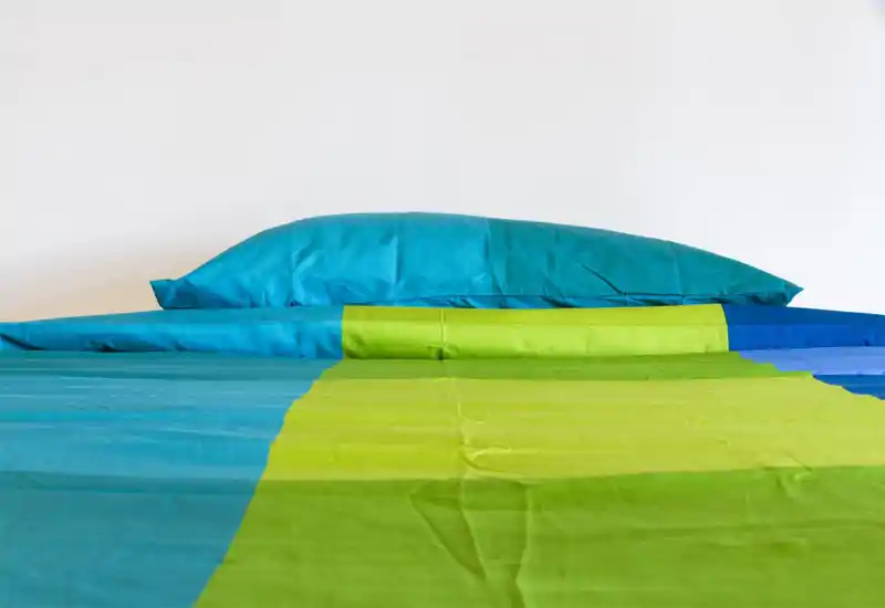 Primo piano di un letto ben rifatto con biancheria turchese e verde lime brillante. L'immagine mette in risalto materasso, cuscino e copriletto colorato in un ambiente minimal e luminoso.