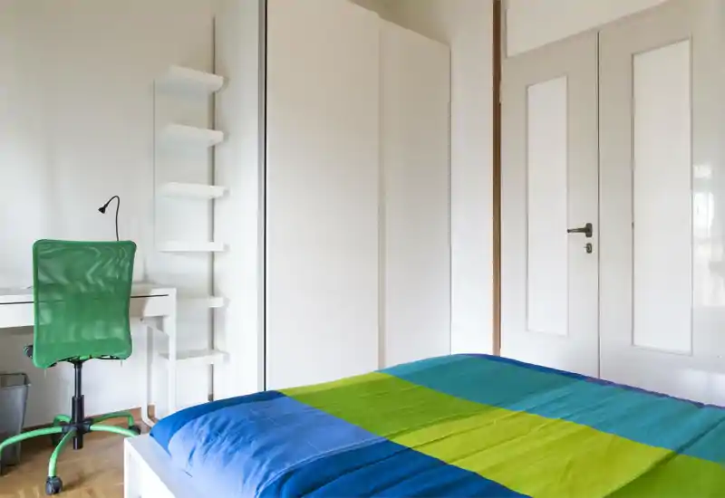 Camera da letto luminosa e minimalista con copriletto blu e verde, armadio bianco e una piccola scrivania con sedia da ufficio verde — spazio pulito e moderno, ideale per una persona.