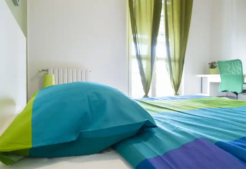 Camera da letto luminosa e colorata con ampio letto in primo piano, luce naturale da grandi finestre e una piccola scrivania sullo sfondo — biancheria blu e verde che crea un’atmosfera vivace.