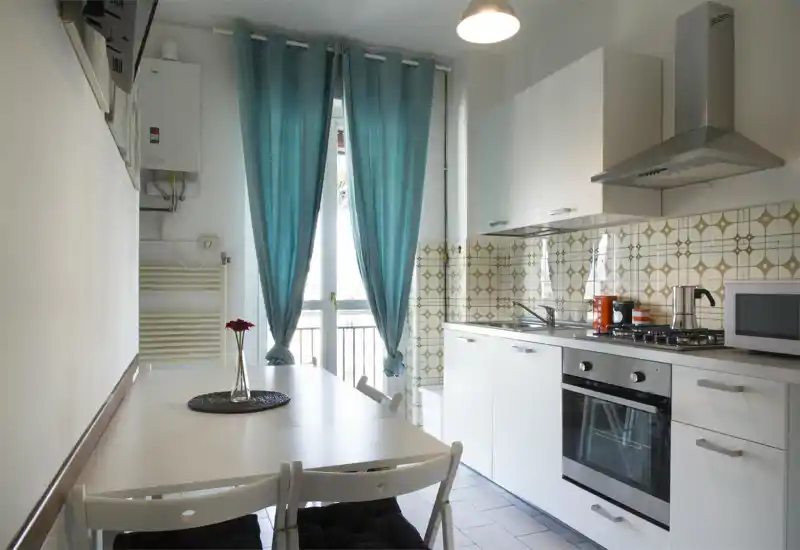 Cucina compatta e luminosa con pensili bianchi, forno incassato e piano cottura a gas, piccolo tavolo da pranzo e tende turchesi che lasciano entrare la luce naturale.