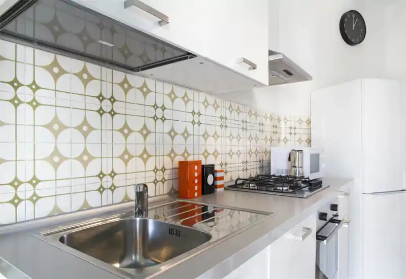 Cucina moderna compatta con lavabo in acciaio inox, piano cottura a gas, pensili bianchi e piastrelle decorative; luminosa e ordinata.