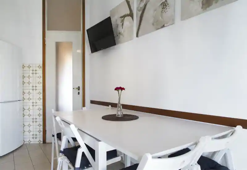 Angolo cucina/pranzo luminoso e compatto con tavolo bianco, sedie pieghevoli con cuscini, quadri decorativi e un vaso con un fiore — ordinato e minimale per appartamenti piccoli.