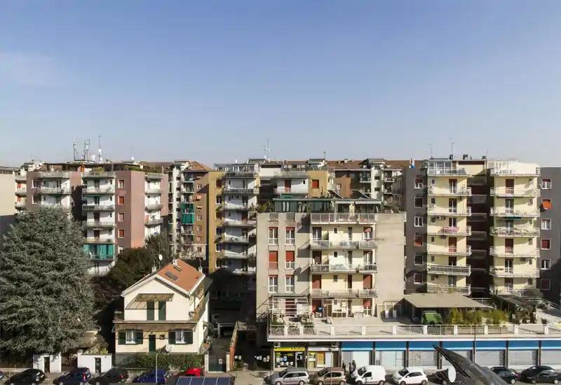 Vista esterna urbana con più condomini dotati di balconi, negozi al piano terra e auto parcheggiate sotto un cielo terso — utile per mostrare la facciata dell’edificio e il quartiere.