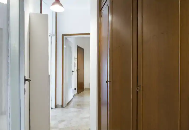 Ingresso luminoso e pulito con armadiature murali in legno, pavimento piastrellato e luce naturale a sinistra; piccolo e ordinato corridoio d'ingresso.