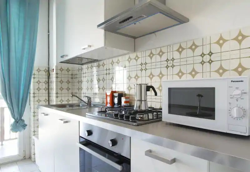 Cucina compatta e luminosa con piano in acciaio, piano cottura a gas, forno incassato e microonde sul piano; piastrelle decorative e tenda azzurra chiara danno carattere.