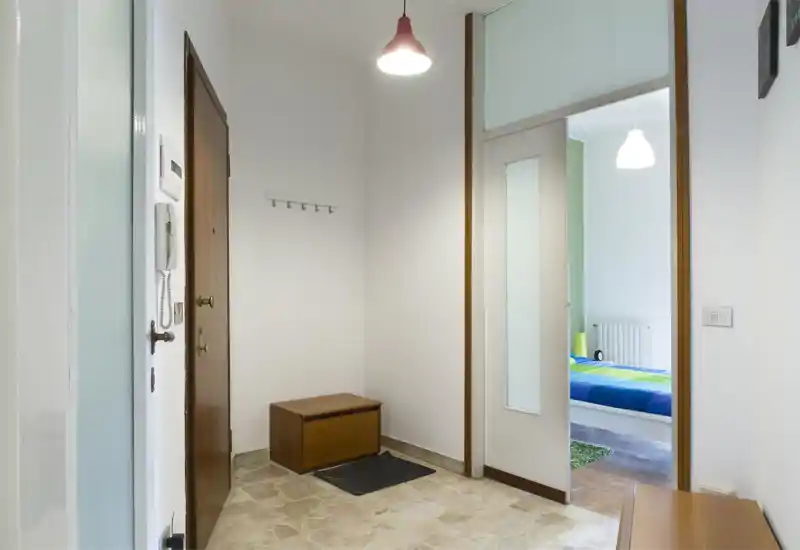 Ingresso luminoso e semplice con porta in legno, citofono, appendiabiti e piccolo panca contenitore — ordine e buona prima impressione.