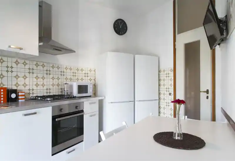 Cucina moderna e luminosa con ante bianche, piano cottura a gas, forno incasso, due frigoriferi e parete piastrellata con motivo; un tavolo con un vaso rende l'ambiente accogliente.