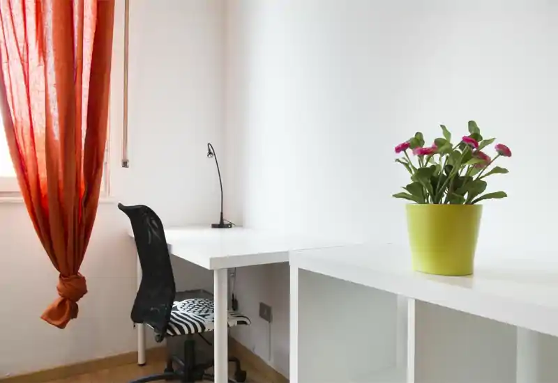 Bright, minimalist home office corner with a white desk, ergonomic mesh chair and a small desk lamp; a green potted plant and warm orange curtain add color.