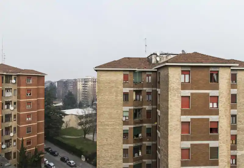 Vista esterna urbana con facciate di condomini, una strada con auto parcheggiate e alberi sotto un cielo coperto.