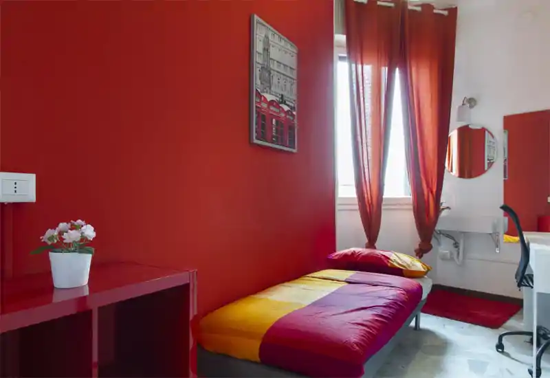 Compact single bedroom with a colorful red-and-yellow bed, matching red accent wall and natural light from a window. The room includes a small desk area and a sink, ideal for a student or short-stay rental.