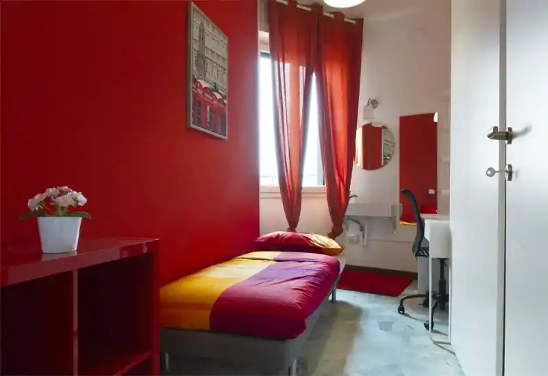 Compact single bedroom with a colorful fitted bed, warm red walls and natural light from the window; includes a small desk and mirror for study or grooming.