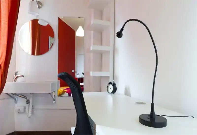 Piccolo angolo studio in camera con scrivania bianca, lampada da tavolo nera flessibile e vista verso l'area letto visibile attraverso la porta; ambiente luminoso e moderno con pareti rosse e mensole minimaliste.