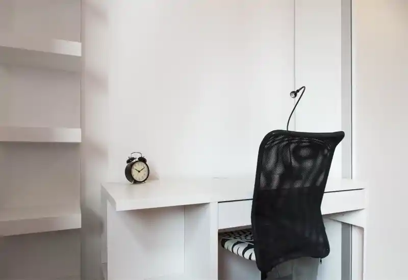 Angolo lavoro minimalista e luminoso con scrivania bianca, sedia da ufficio con schienale in rete nero, piccola lampada da lavoro e una sveglia analogica: un angolo studio moderno e pulito.