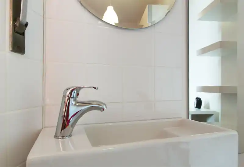 Primo piano di un lavabo da bagno con rubinetto cromato su parete piastrellata bianca; zona sanitaria pulita, essenziale e ben illuminata.