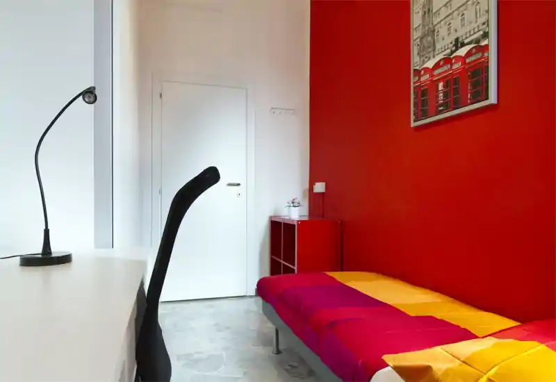 Small, well-lit bedroom with a single bed dressed in bright bedding, a minimalist desk and chair, and bold red accent wall with artwork.