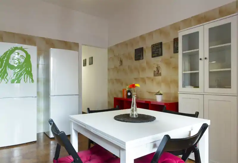 Bright, stylish dining area with a white table, black folding chairs with red cushions, decorative vase centerpiece and a cabinet; warm tiled walls and two refrigerators visible in the background.