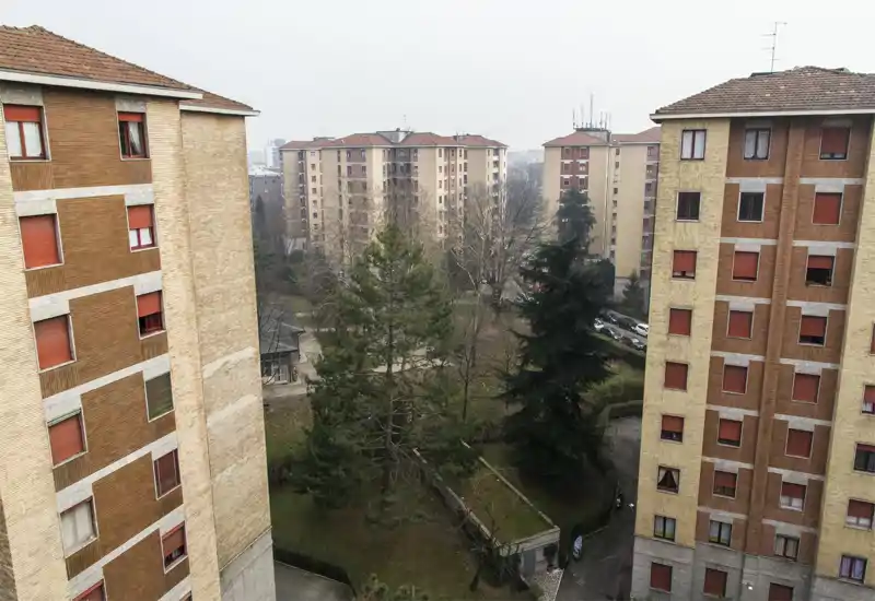 Exterior view of a residential courtyard and neighboring apartment blocks seen from a higher-floor window or balcony; mature trees and pathways are visible beneath an overcast sky.