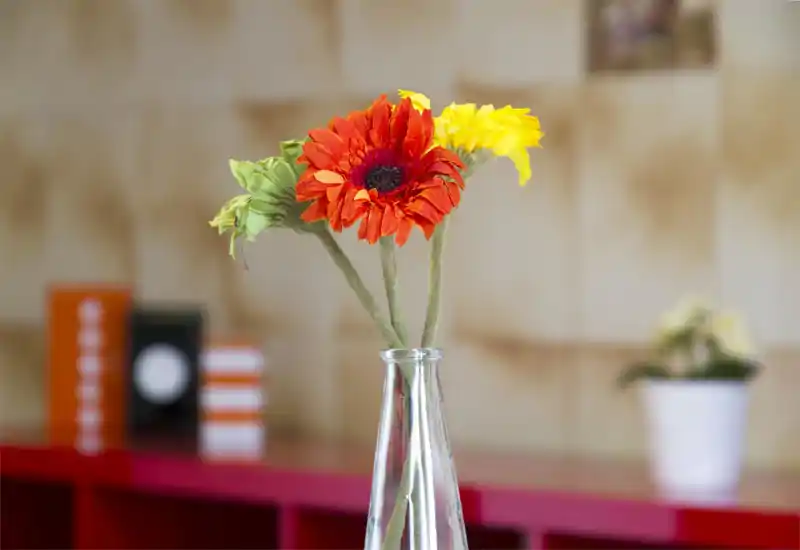 Close-up of a decorative flower arrangement in a glass vase on a table, with a softly blurred living area background — a small detail shot that highlights homey décor.