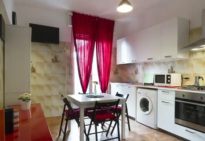 Bright fitted kitchen with white cabinets, built-in oven, washing machine and a small dining table with four chairs; bold red curtains and cushions add color and warmth.